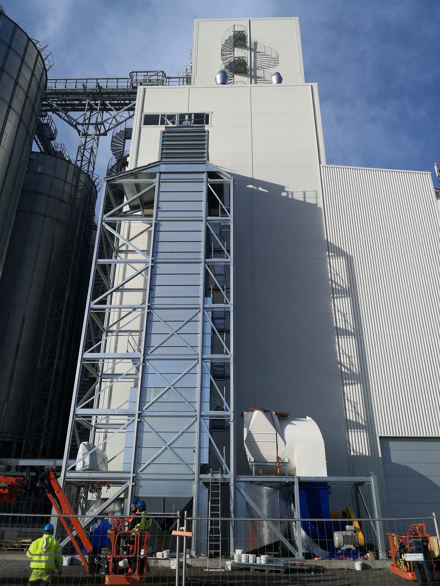 Wysoka hala przemysłowa z metalową konstrukcją i spiralnymi schodami na dachu. Widoczni pracownicy na podnośniku koszowym podczas montażu elementów.