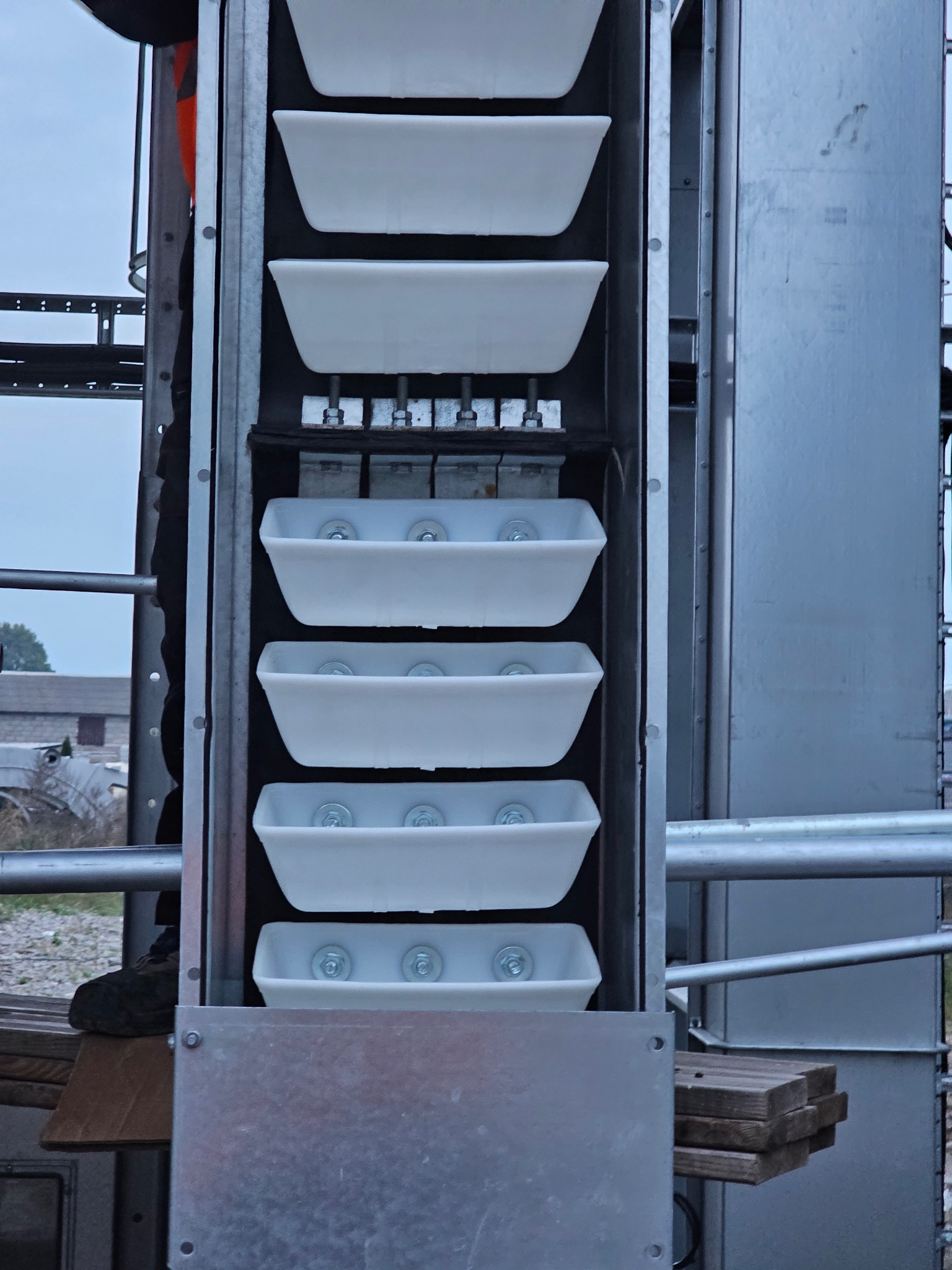 Metalowa konstrukcja hali z rzędami białych pojemników, widoczne elementy montażowe i fragment otoczenia zewnętrznego. Detal konstrukcyjny w surowym oświetleniu.