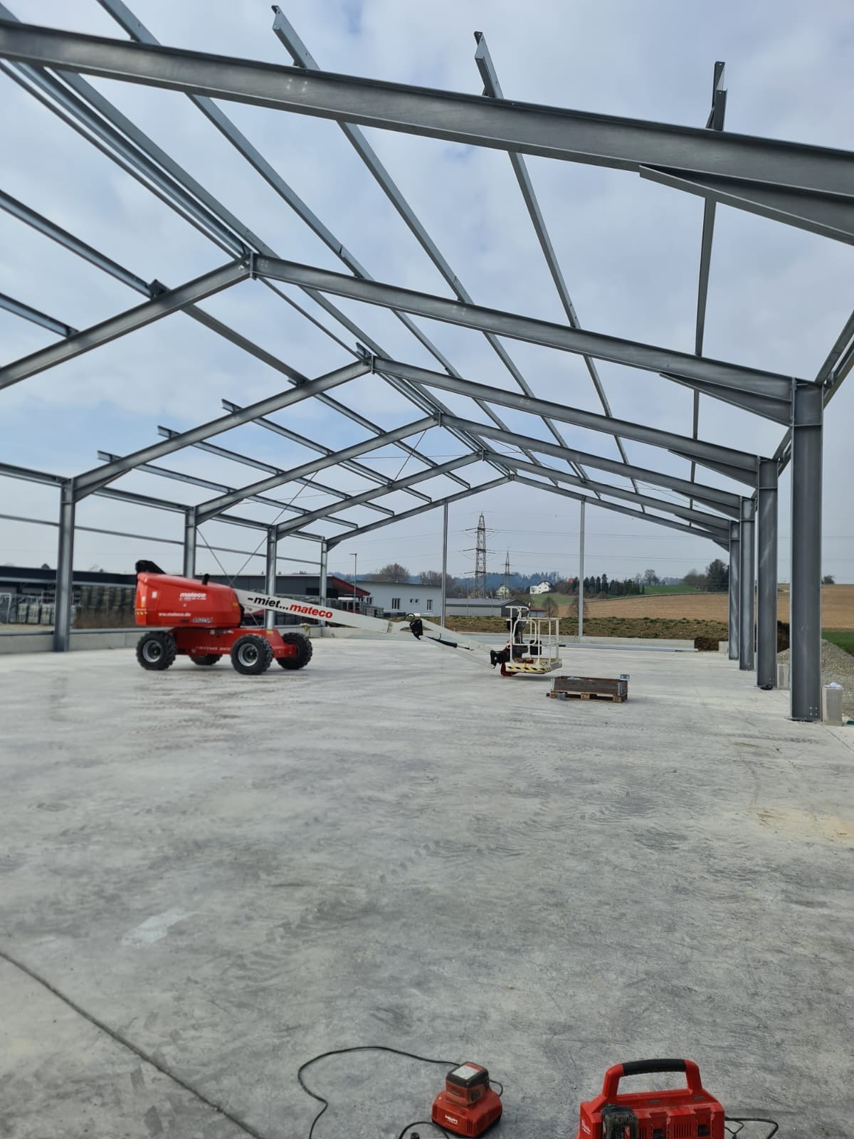 Konstrukcja stalowa hali w trakcie budowy, widoczny podnośnik koszowy i narzędzia na betonowej posadzce. Słupy i kratownica tworzą szkielet budynku.