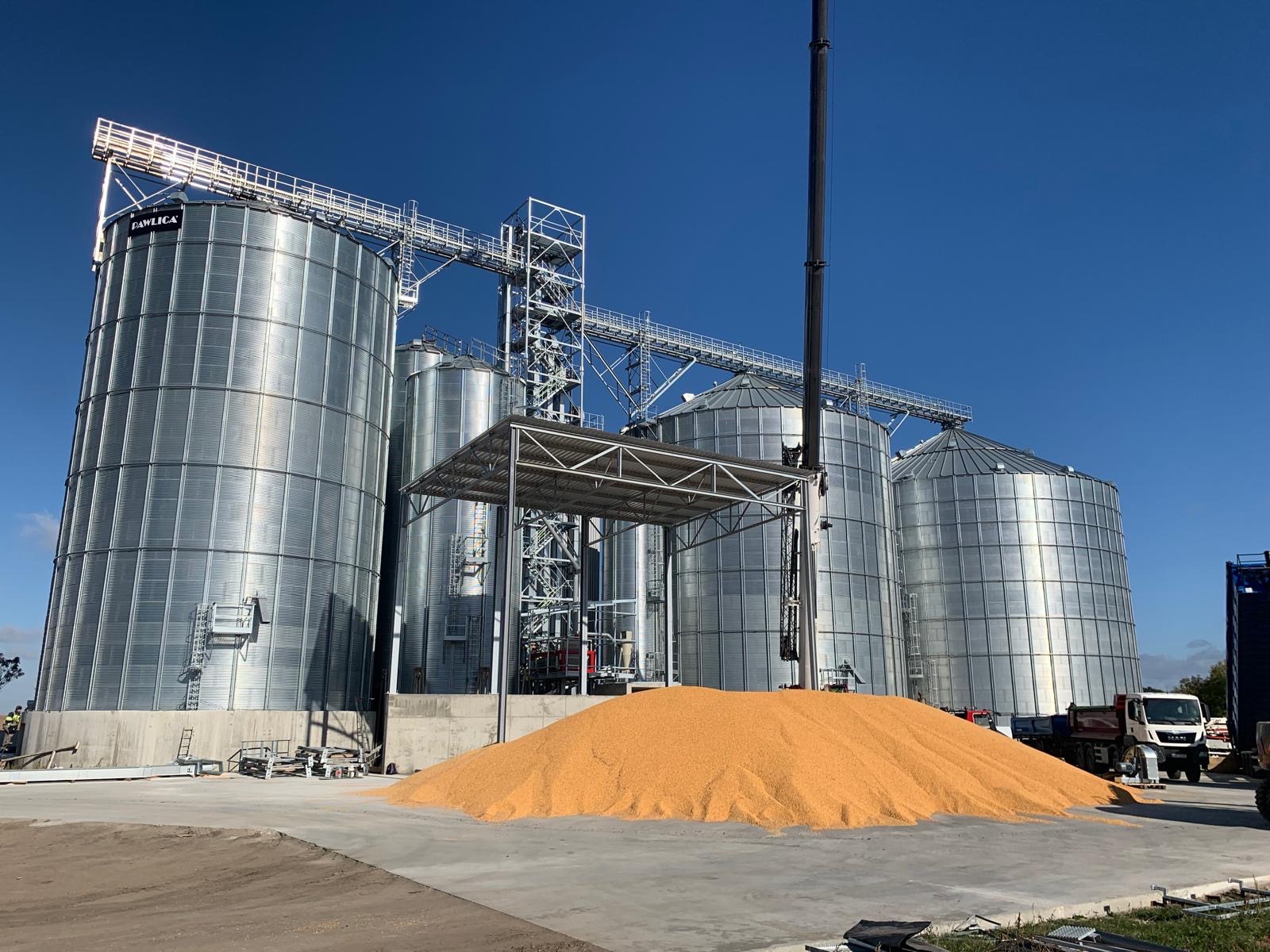 Kompleks silosów zbożowych ze stali nierdzewnej w słoneczny dzień, z górą ziarna u podstawy. Widoczna konstrukcja wsporcza i ciężarówka w tle.