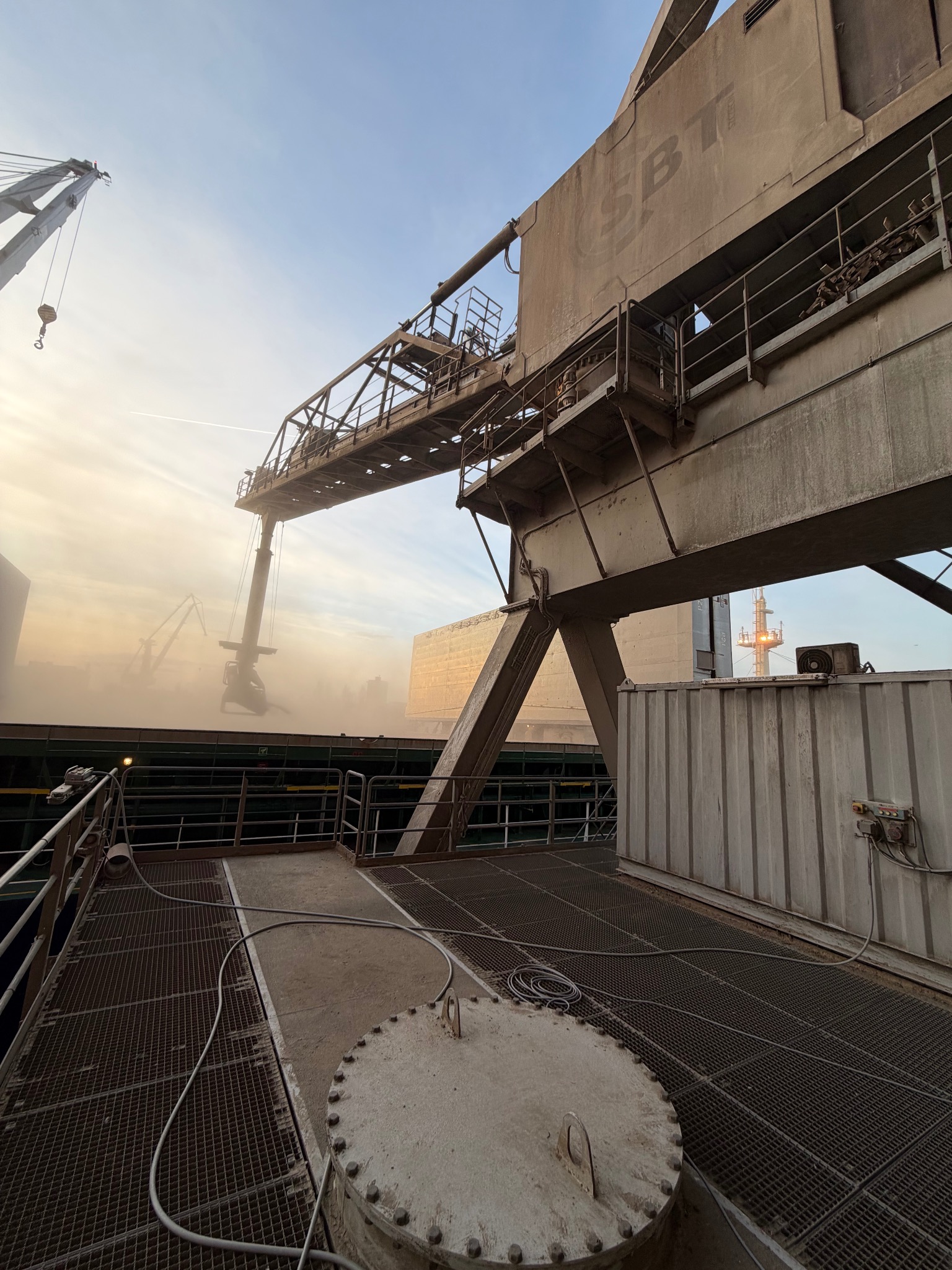 Widok na suwnicę portową SBT w trakcie załadunku, z widoczną konstrukcją stalową i elementami infrastruktury przemysłowej w tle, w warunkach zapylenia.