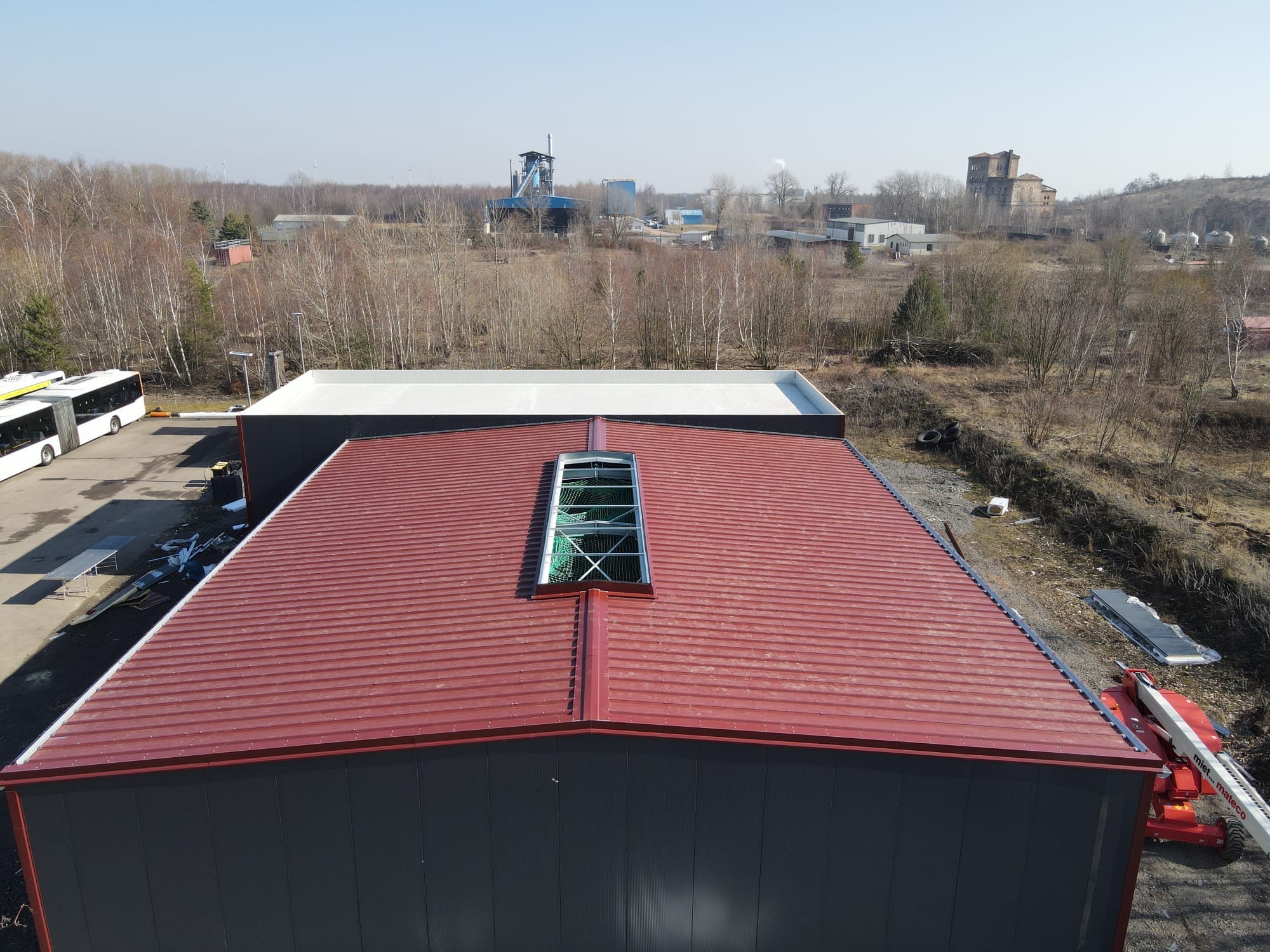 Hala z bordową blachą trapezową i świetlikiem dachowym, widok z góry. W tle zabudowania przemysłowe i las. Na placu zaparkowane autobusy i podnośnik koszowy.
