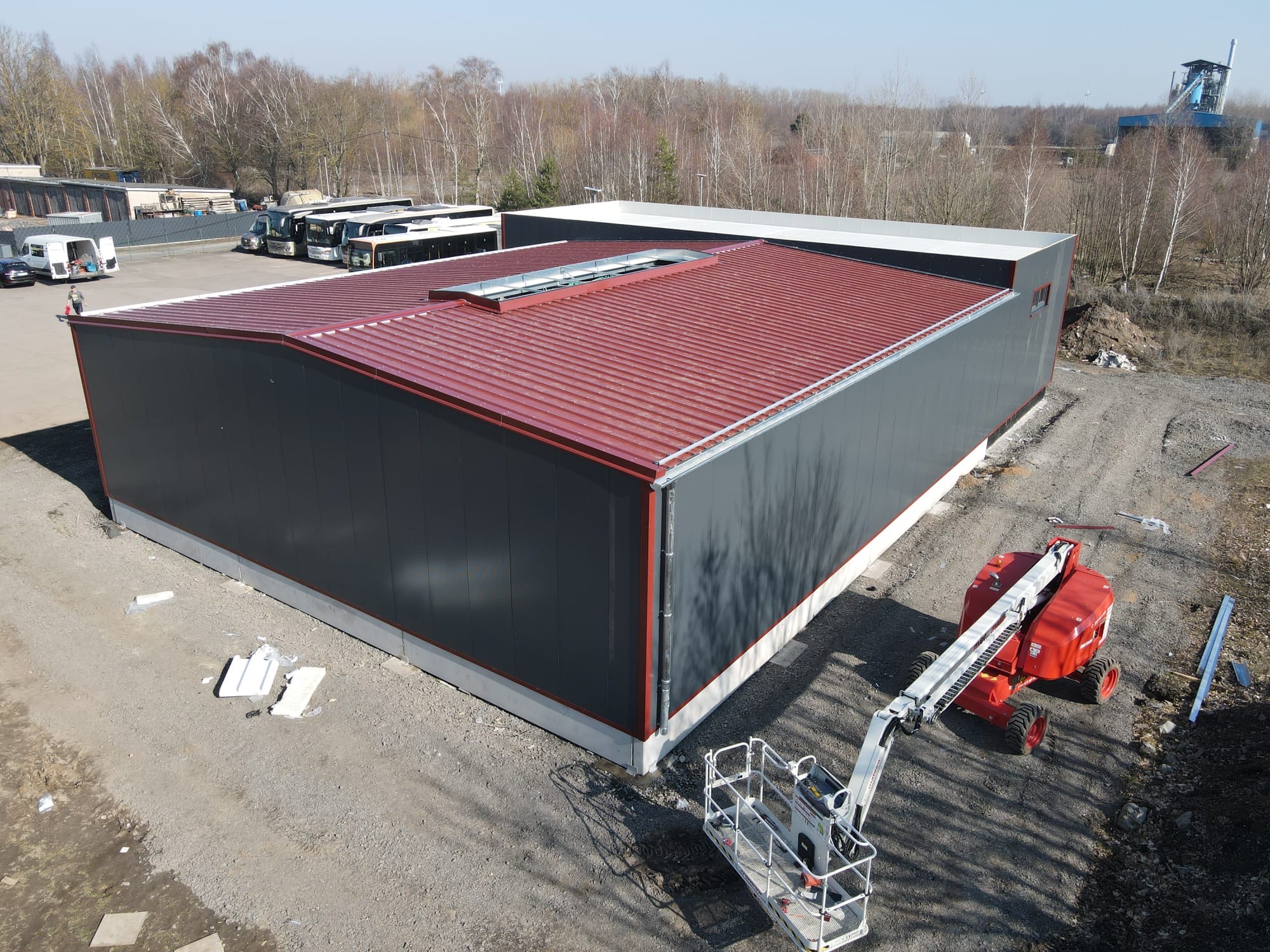 Nowoczesna hala z szarymi ścianami i bordowym dachem z blachy trapezowej, widok z góry, na placu budowy z podnośnikiem koszowym i zaparkowanymi autobusami w tle.