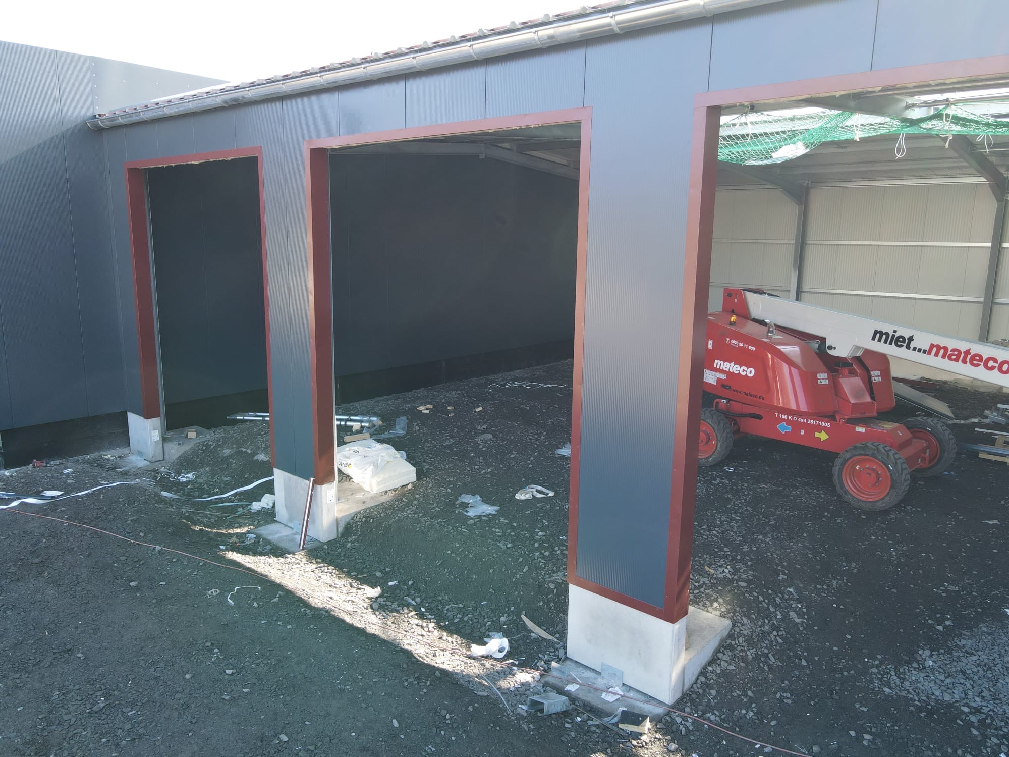 Wznoszenie hali z płyt warstwowych: widok na konstrukcję z trzema otworami wejściowymi, podnośnik koszowy Mateco w tle, teren budowy z kablami i gruzem.