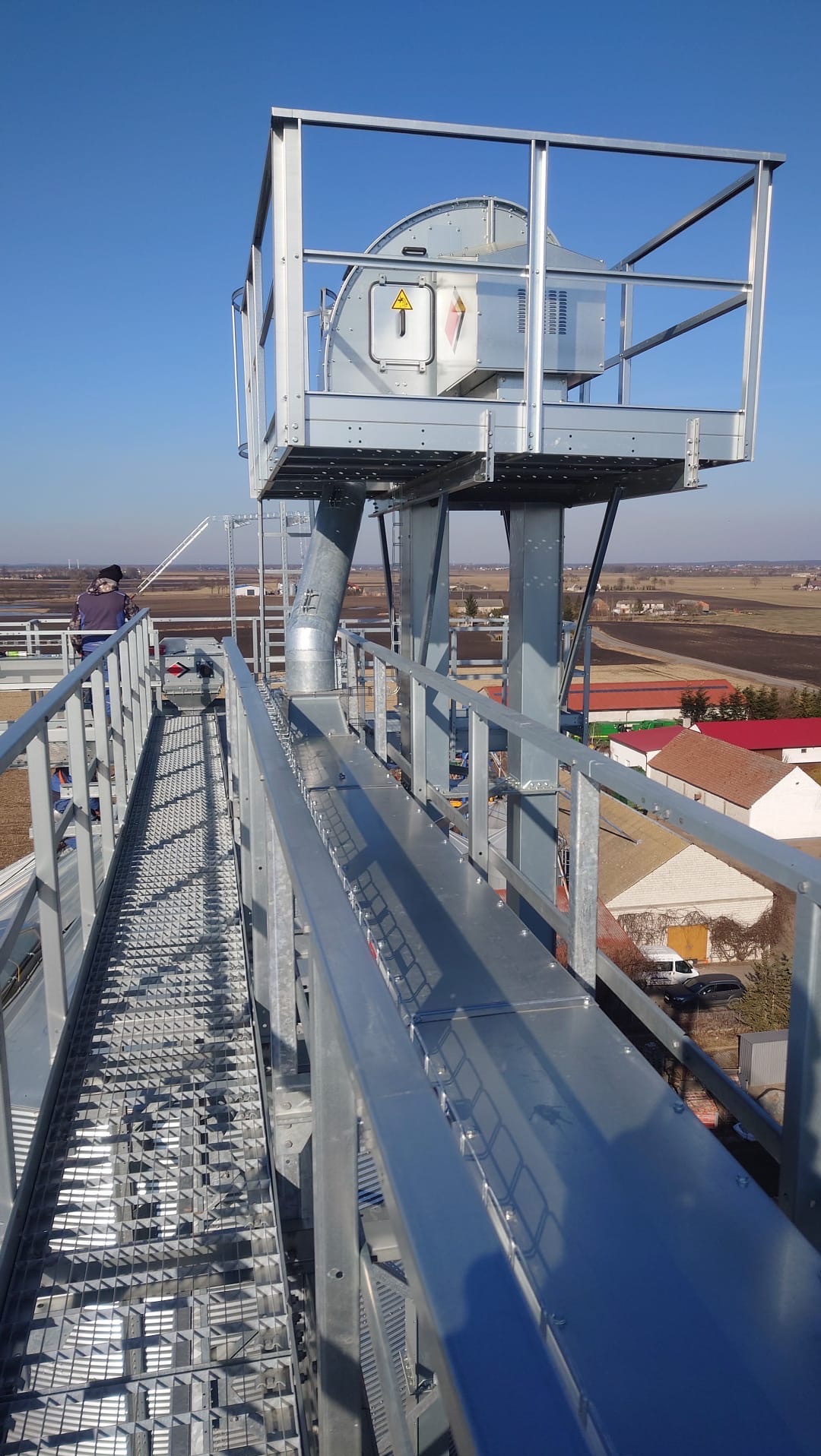 Stalowa konstrukcja hali z platformą i rurą, widok z góry na tle krajobrazu. Metalowa kładka z kraty, barierki. Osoba w oddali na platformie.