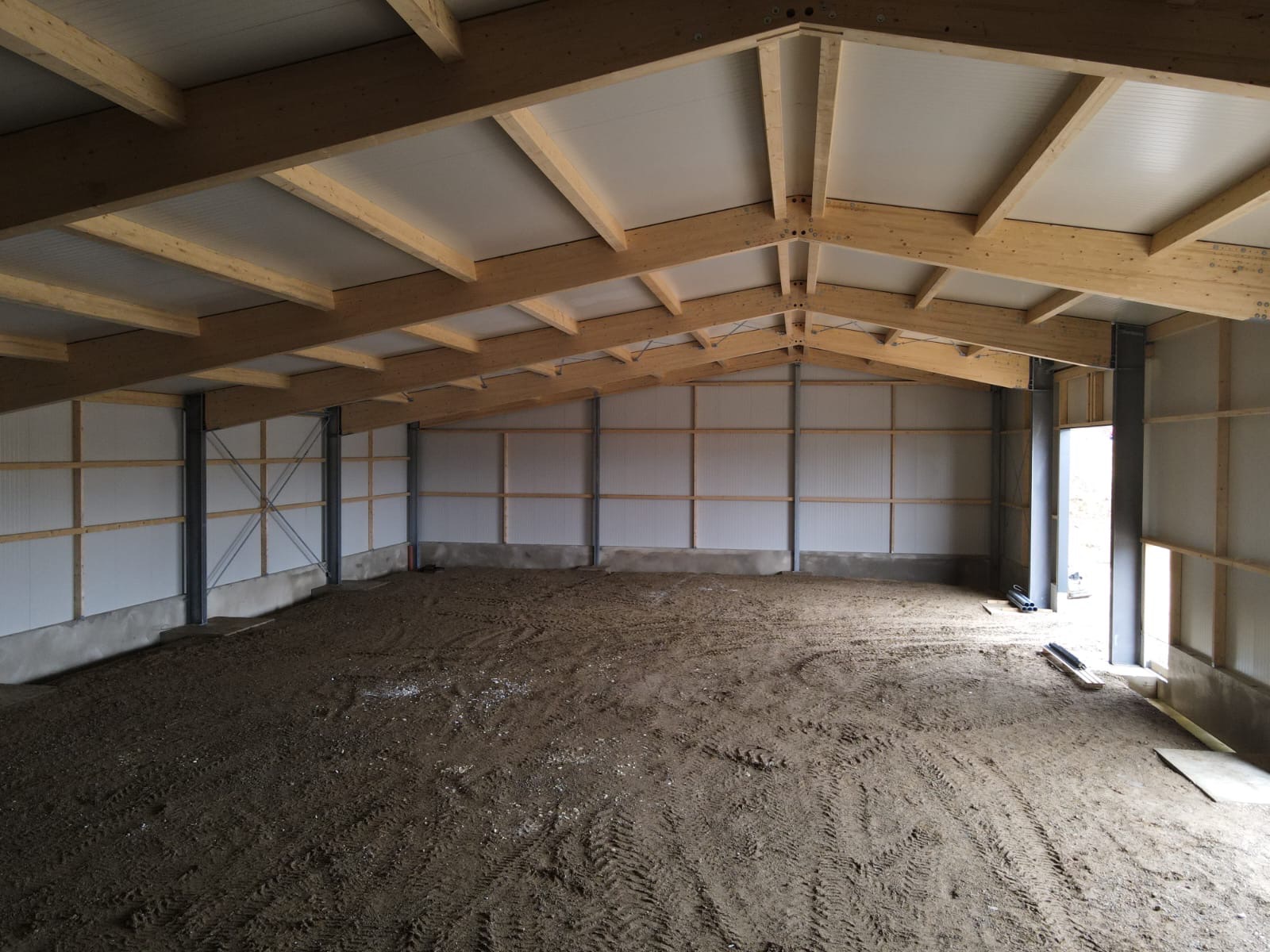 Wnętrze hali w trakcie budowy: widoczne drewniane elementy konstrukcyjne dachu, ściany z płyt warstwowych i surowa podłoga z ubitej ziemi. Stalowe słupy podtrzymują konstrukcję.