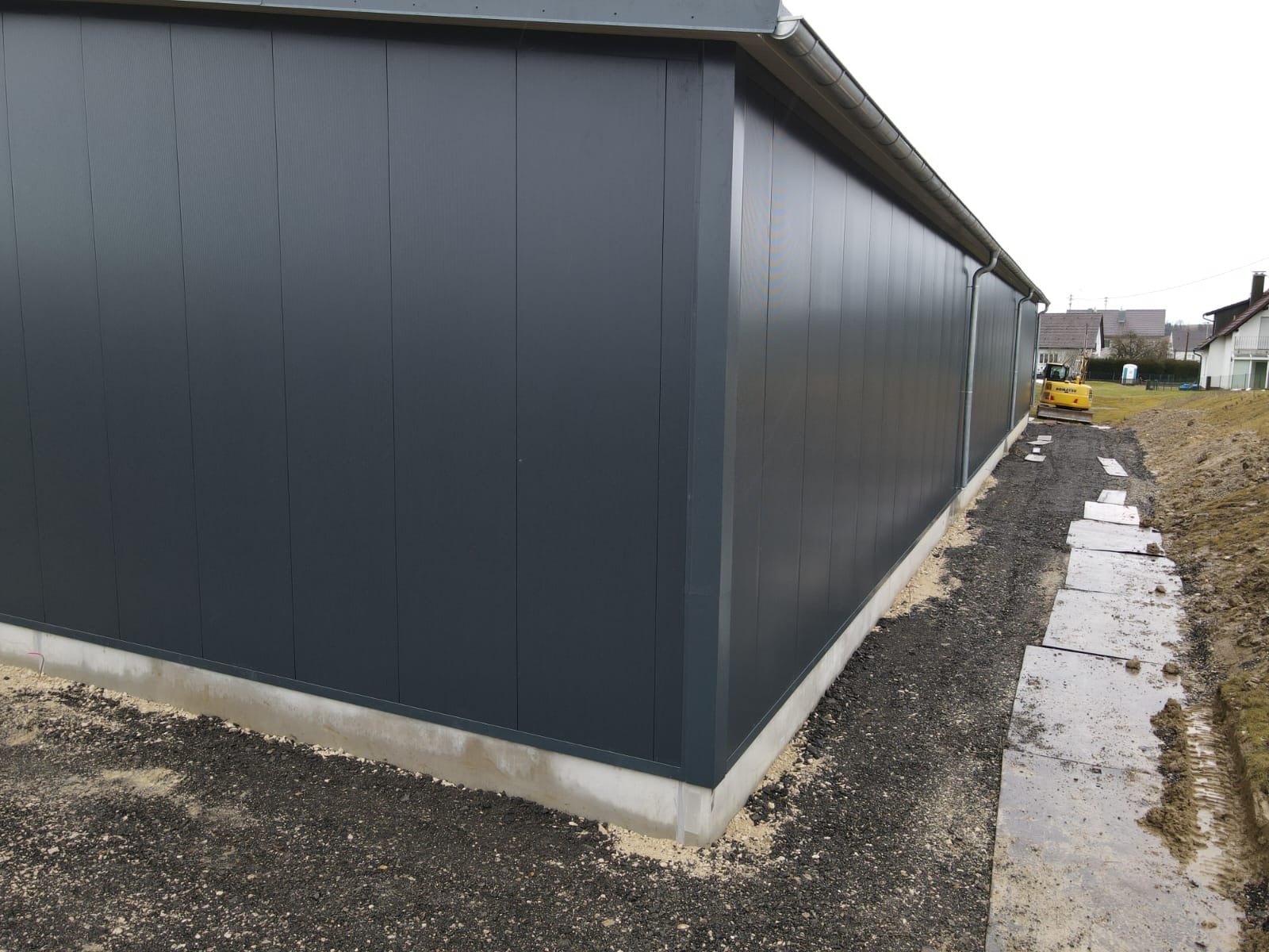 Szara hala z płyt warstwowych, widok z zewnątrz, zarys fundamentu, rynna. W tle żółta koparka i zabudowa jednorodzinna. Ujęcie z dołu, podkreślające wysokość konstrukcji.