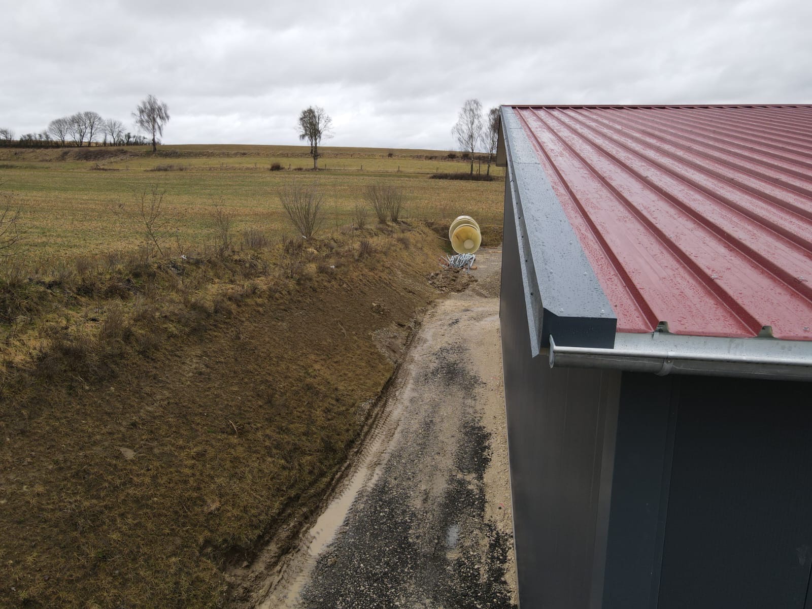 Czerwony dach hali z blachy trapezowej, szara ściana, rynna. W tle pole i drzewa pod pochmurnym niebem. Widoczne materiały budowlane obok hali.
