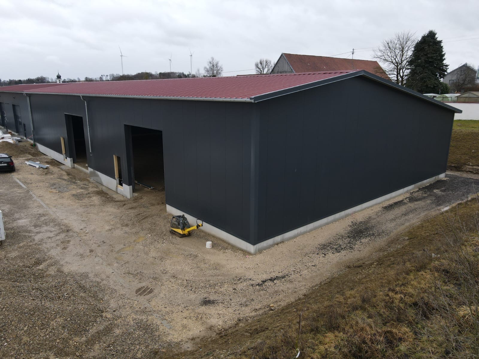 Gotowa hala z ciemnoszarą elewacją z płyt warstwowych i czerwonym dachem, widok z góry, na placu budowy stoi mała zagęszczarka.