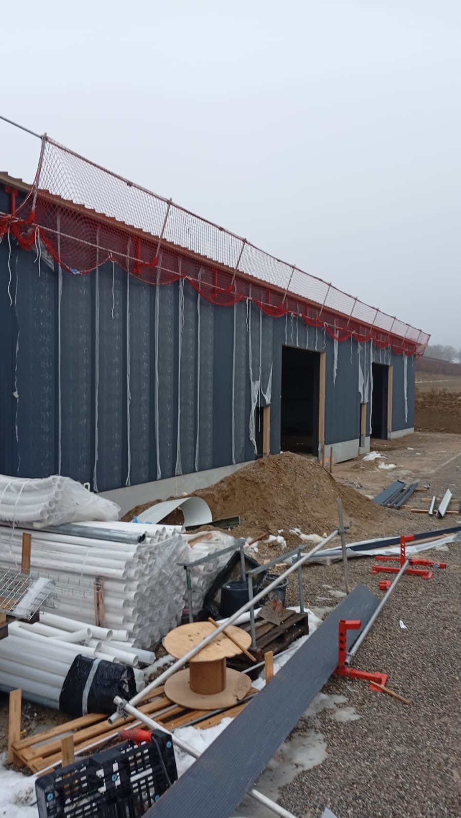 Budowa hali w trakcie montażu płyt warstwowych. Widoczne elementy konstrukcyjne, materiały budowlane i siatka zabezpieczająca na krawędzi dachu. Pochmurny dzień.