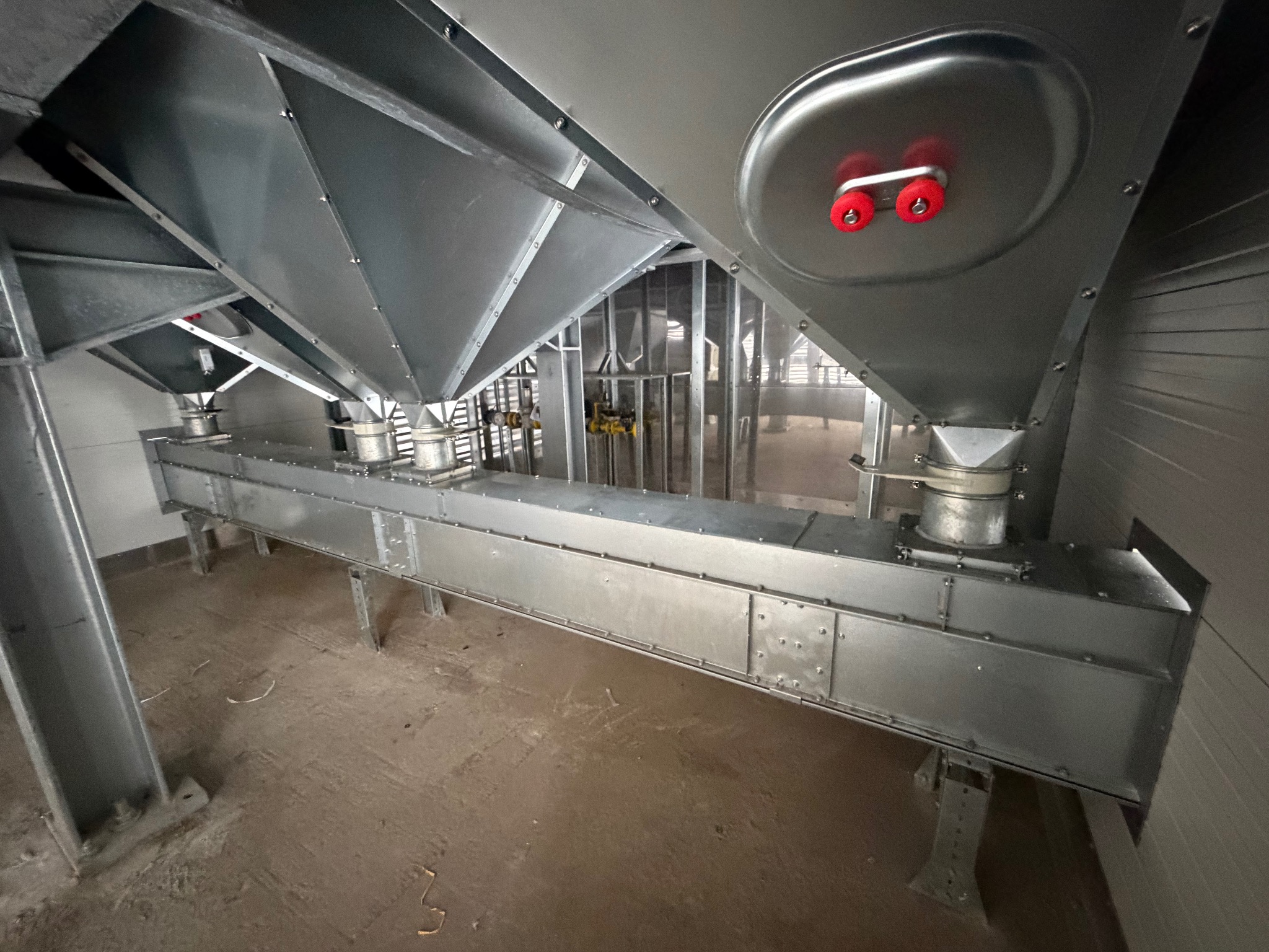 Wnętrze hali z elementami konstrukcji stalowej: metalowe silosy z czerwonymi rolkami, połączone poziomym kanałem. Widoczne elementy mocujące i wsporniki.