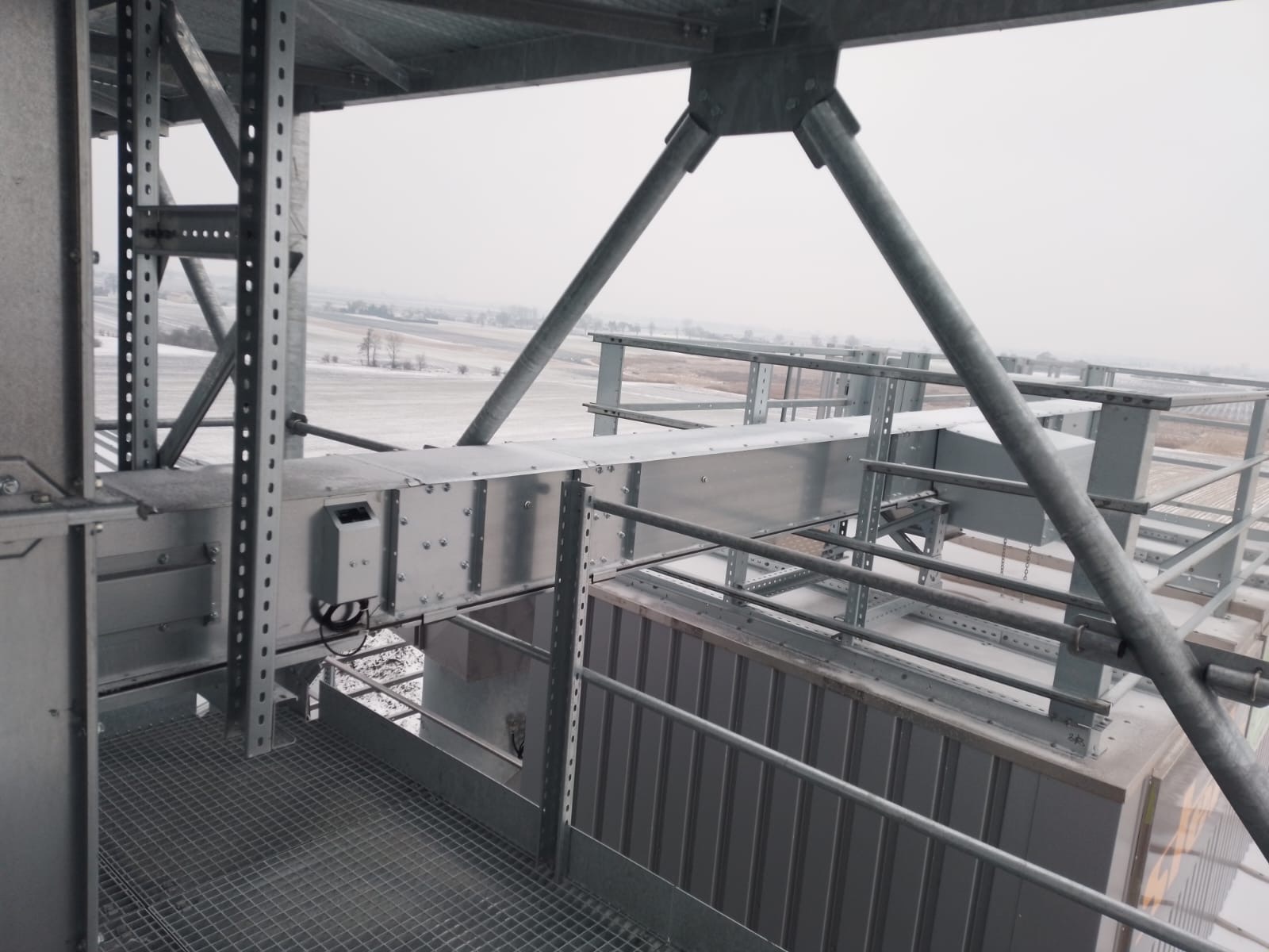 Wnętrze stalowej konstrukcji hali z widokiem na zimowy krajobraz. Metalowe elementy połączone śrubami, kratownica podłogowa, szare tło. Detal konstrukcyjny.