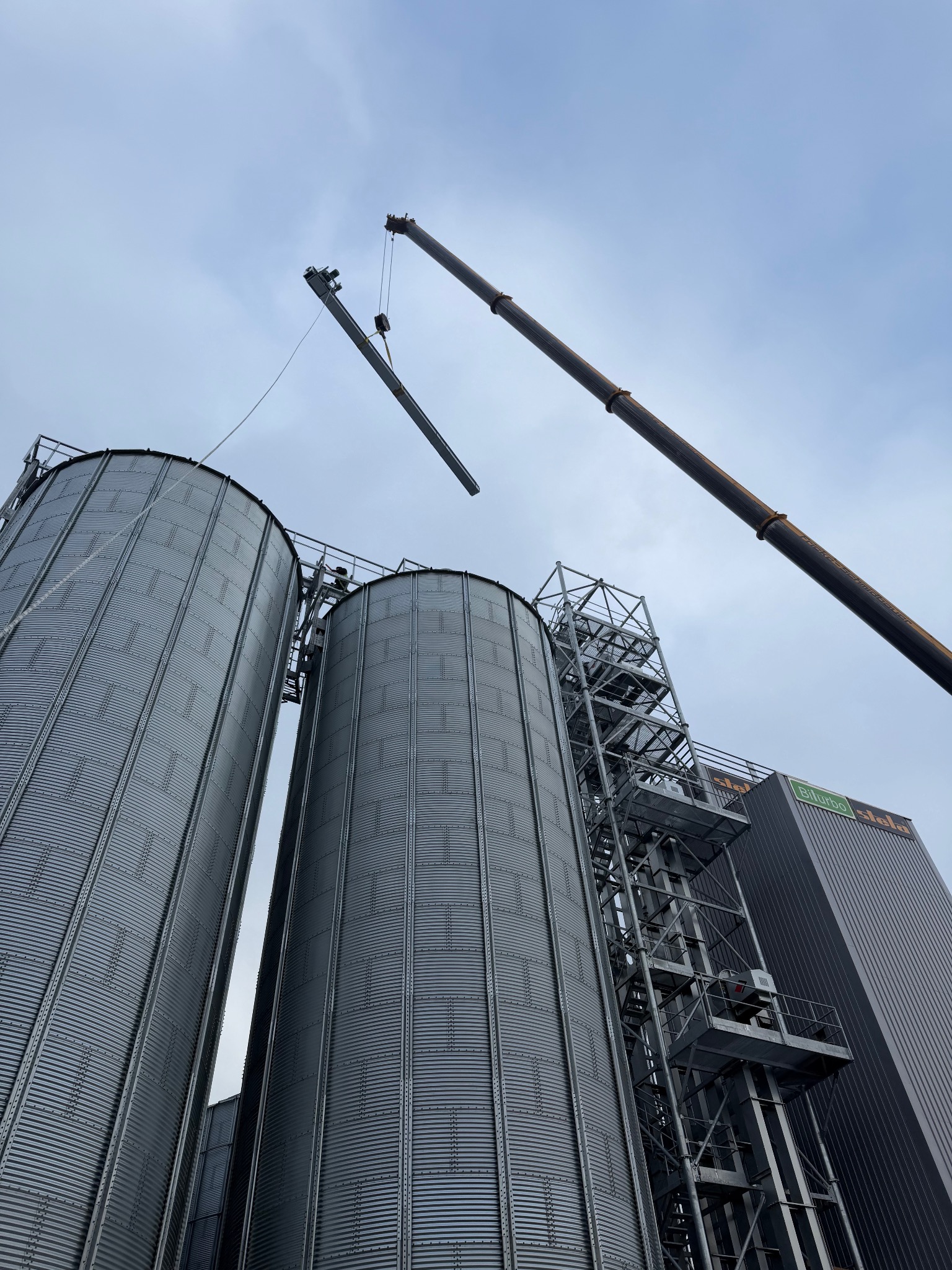 Montaż dwóch srebrnych silosów z użyciem dźwigu na tle pochmurnego nieba. Widok z dołu podkreśla skalę konstrukcji i etap budowy.