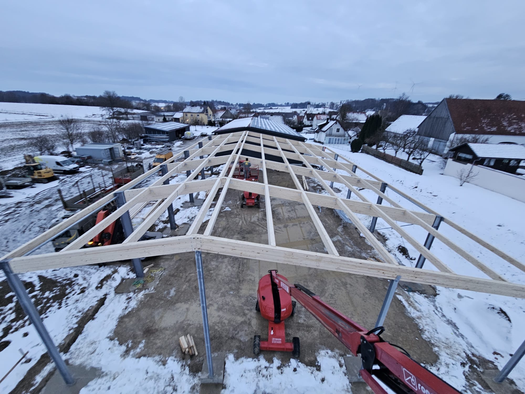 Konstrukcja hali w zimowym krajobrazie. Widok z góry na drewniany szkielet hali z metalowymi słupami, z użyciem podnośników. W tle ośnieżone pola i zabudowania.