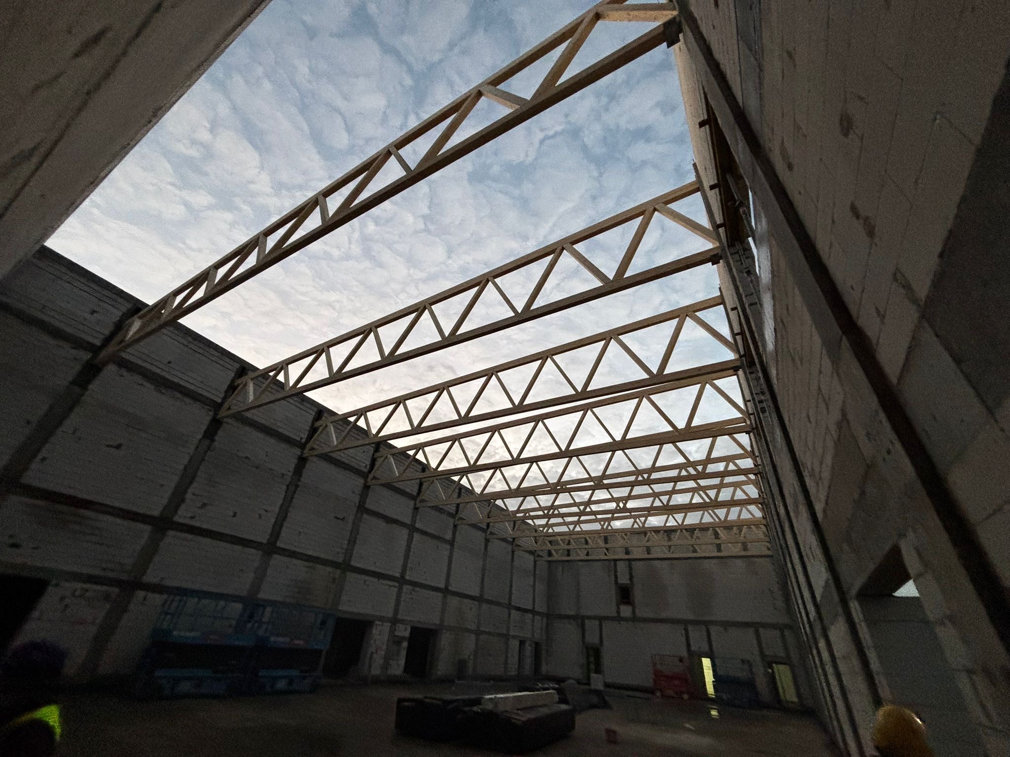 Wnętrze hali w budowie z widoczną konstrukcją dachu z drewnianych belek i fragmentem nieba. Ściany z betonu komórkowego, widoczne okna i sprzęt budowlany.