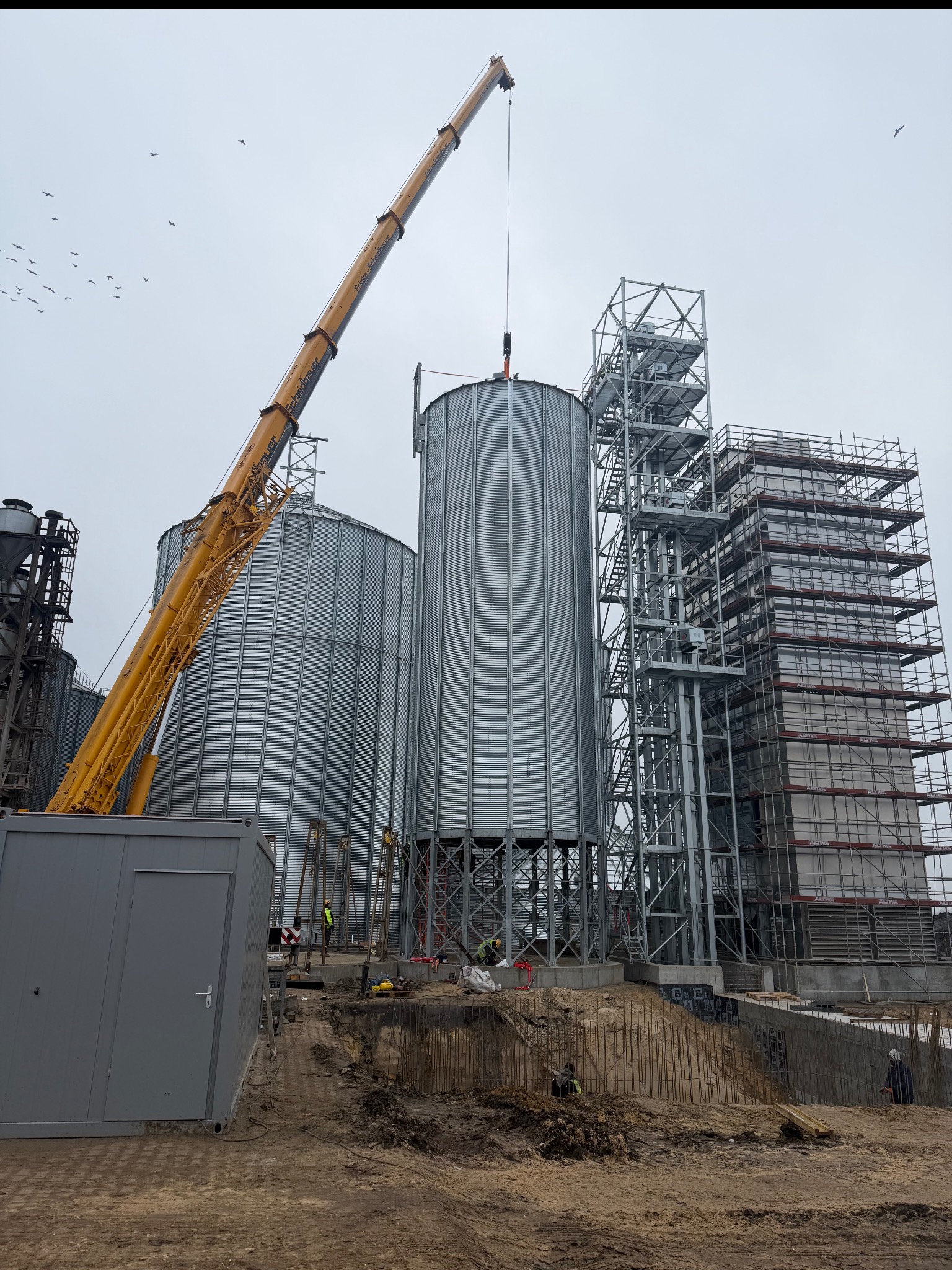 Budowa kompleksu silosów zbożowych z użyciem dźwigu, widoczne stalowe konstrukcje, szalunki i pracownicy w odblaskowych kamizelkach na placu budowy.