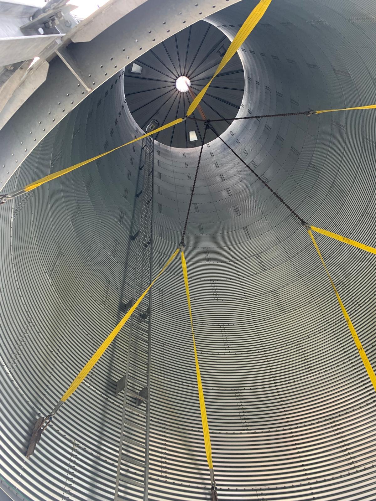 Wnętrze stalowej hali z widoczną drabiną i żółtymi pasami mocującymi, perspektywa od dołu, okrągły otwór w górnej części konstrukcji.