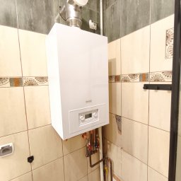 Kamil Góralski - Biały piec gazowy z chromowanym kominem w łazience z beżowymi i szarymi płytkami. Widoczne rury i zawory instalacji. Ściana z uszkodzonym fragmentem przyłącza.