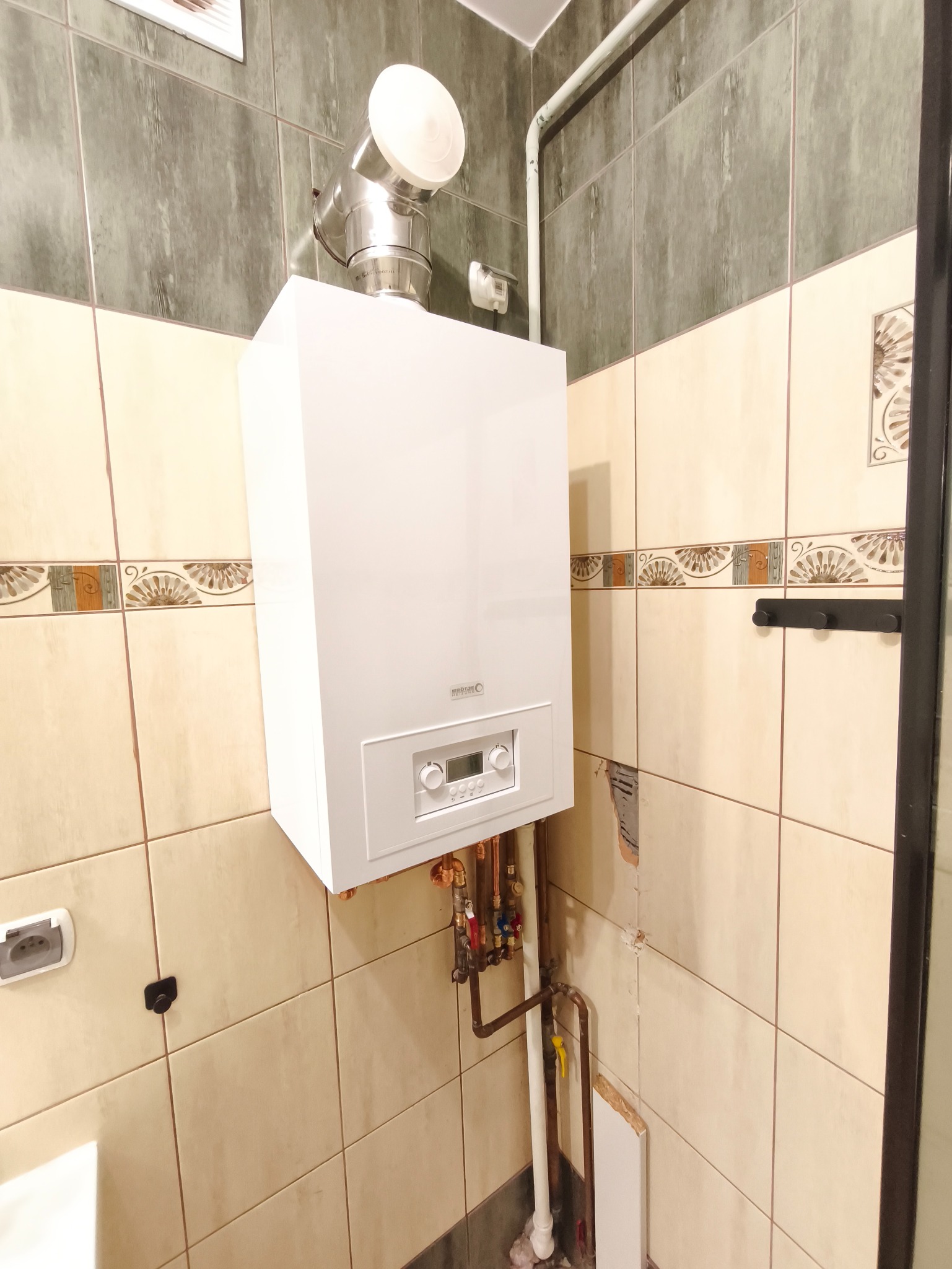Biały piec gazowy z chromowanym kominem w łazience z beżowymi i szarymi płytkami. Widoczne rury i zawory instalacji. Ściana z uszkodzonym fragmentem przyłącza.