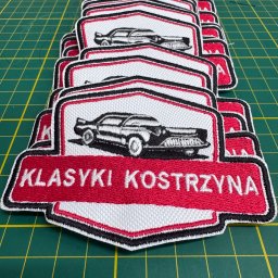 Waszehafty.pl Anna Stasiw - Stos haftowanych naszywek 'Klasyki Kostrzyna' z motywem czarnego samochodu na białym tle z czerwonym napisem, leżących na zielonej macie do cięcia.