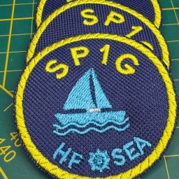 Waszehafty.pl Anna Stasiw - Stos trzech okrągłych naszywek z motywem żaglówki i napisem 'SP1G HF SEA', obramowanych żółtą nicią, leżących na zielonej macie do cięcia.