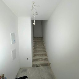 JMP Rzeszów - Wnętrze w trakcie remontu: białe ściany, surowe schody betonowe, wisząca żarówka i narzędzia malarskie. Widoczne puszki elektryczne i instalacja. Minimalistyczny kadr.