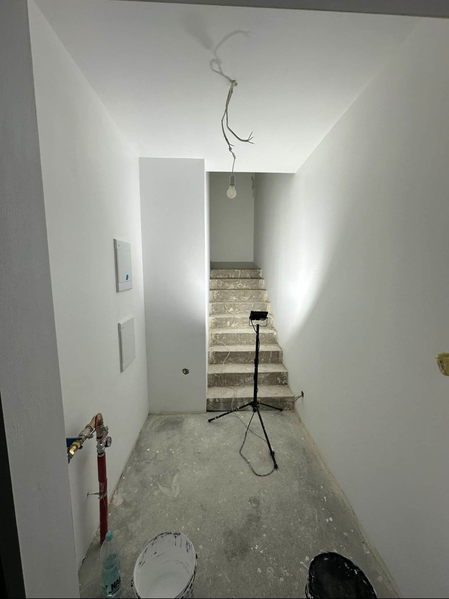 Wnętrze po malowaniu: białe ściany, schody w tle, widoczny sprzęt oświetleniowy i puszki z farbą. Minimalistyczny styl, surowe wykończenie.