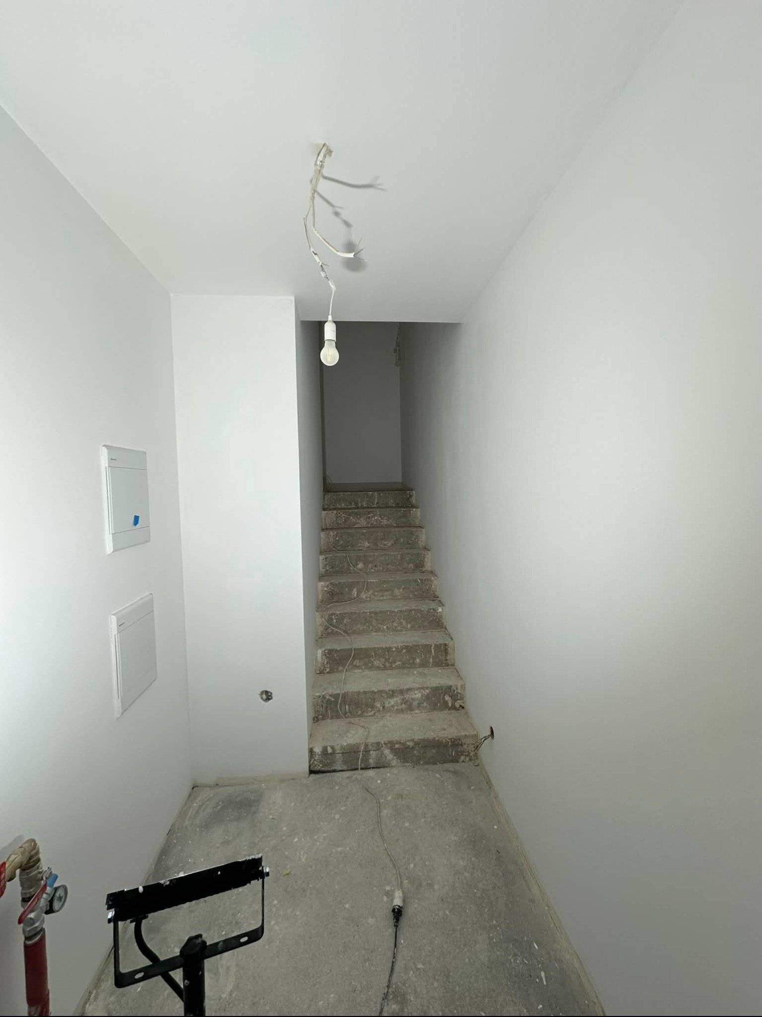 Wnętrze w trakcie remontu: białe ściany, surowe schody betonowe, wisząca żarówka i narzędzia malarskie. Widoczne puszki elektryczne i instalacja. Minimalistyczny kadr.