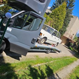 Kacpi - Szary Iveco laweta z białym Fiatem 125p na platformie, zabezpieczonym pasami transportowymi. W tle budynek i zieleń. Ujęcie z dołu, lekko obrócone.