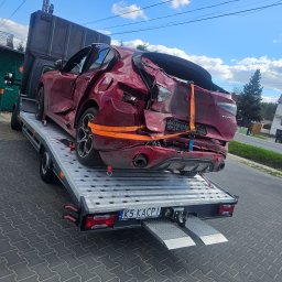 Kacpi - Laweta z uszkodzonym, czerwonym samochodem Alfa Romeo Stelvio, zabezpieczonym pomarańczowymi pasami transportowymi. Widoczne tablice rejestracyjne lawety.