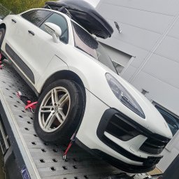 Kacpi - Biały samochód osobowy marki Porsche z bagażnikiem dachowym, zabezpieczony pasami, umieszczony na platformie lawety. Widok z boku, lekki kąt.