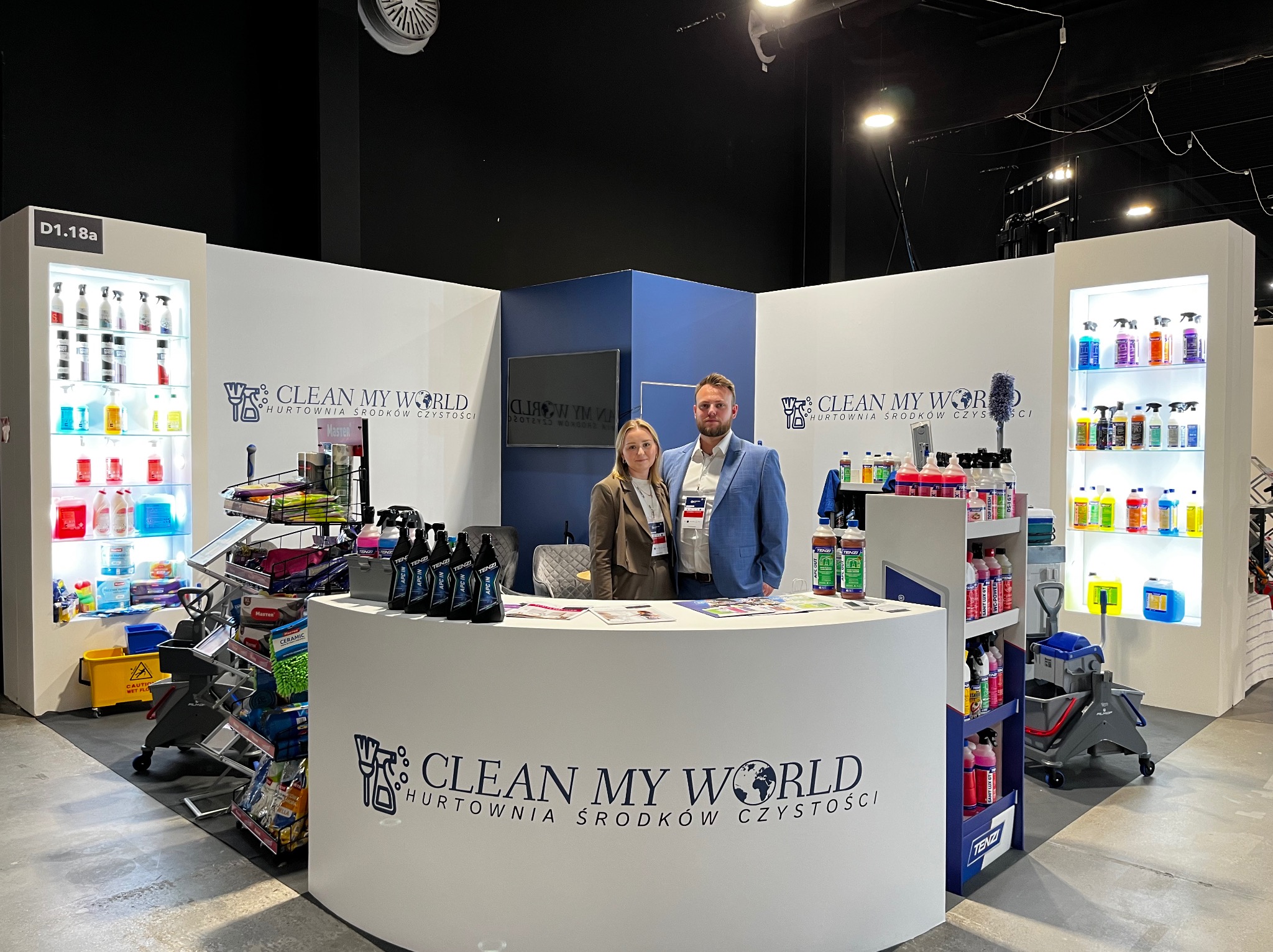 Stoisko targowe firmy Clean My World z ekspozycją środków czystości, mopów i wózków. Dwie osoby za ladą. Targi Warsaw CleanTech Expo. Oświetlenie punktowe.
