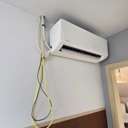 HYDROS MARKET BUDOWLANY SPÓŁKA Z OGRANICZONĄ ODPOWIEDZIALNOŚCIĄ - Biały klimatyzator Daikin zamontowany na ścianie, widoczne przewody w peszlu i luźno wiszące kable. U dołu brązowa ściana z włącznikami. Fragment futryny drzwi po prawej.