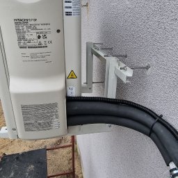 HYDROS MARKET BUDOWLANY SPÓŁKA Z OGRANICZONĄ ODPOWIEDZIALNOŚCIĄ - Zewnętrzna jednostka pompy ciepła Hitachi zamontowana na ścianie budynku. Widoczne rury instalacyjne i wsporniki montażowe. Detal montażu.