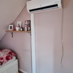 HYDROS MARKET BUDOWLANY SPÓŁKA Z OGRANICZONĄ ODPOWIEDZIALNOŚCIĄ - Biały klimatyzator LG Dual Inverter zamontowany na ścianie w pokoju na poddaszu. Widoczne kable. Na ścianie półka z dekoracjami. Drewniana podłoga.