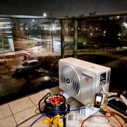 HYDROS MARKET BUDOWLANY SPÓŁKA Z OGRANICZONĄ ODPOWIEDZIALNOŚCIĄ - Biała jednostka zewnętrzna klimatyzacji Kaisai na balkonie, podłączona do narzędzi serwisowych. Widok na nocny, deszczowy krajobraz miejski za szklaną balustradą.