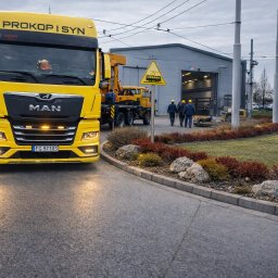 PROKOP I SYN - MICHAŁ PROKOP - Żółta ciężarówka MAN z napisem 'PROKOP I SYN' na tle szarego budynku i grupy robotników w kaskach. Widoczny znak ostrzegawczy i elementy krajobrazu.