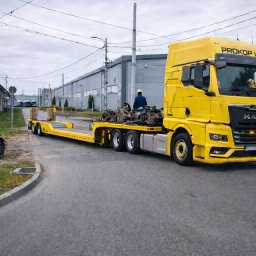 PROKOP I SYN - MICHAŁ PROKOP - Żółty ciągnik siodłowy z naczepą niskopodwoziową przewozi elementy maszyn budowlanych na tle budynków przemysłowych. Widoczny operator w kasku.