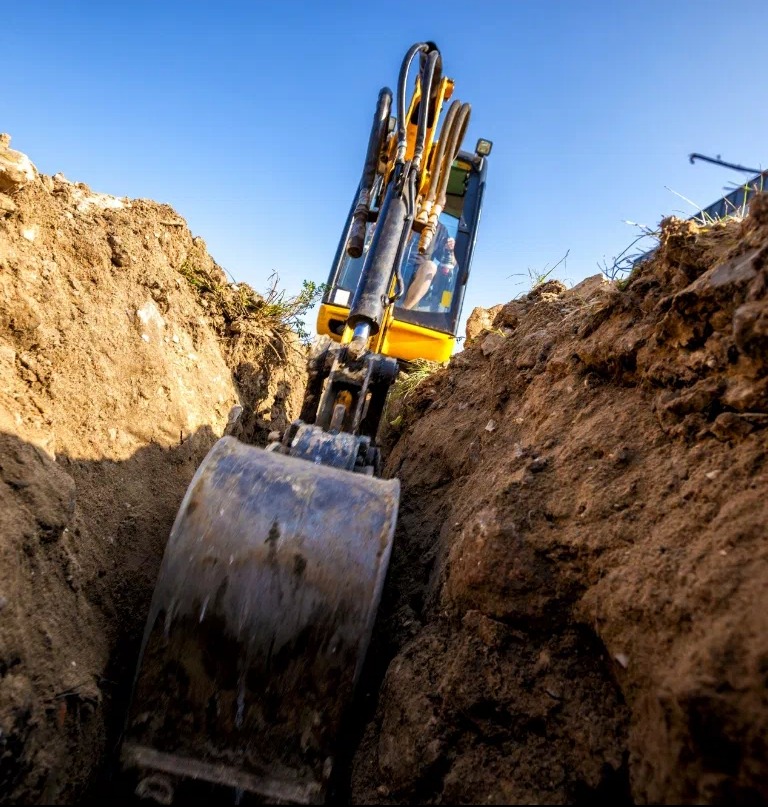 Żółta koparka w trakcie wykopu wąskiego rowu w Karsko, widok z dołu. Maszyna zagłębia się w ziemi, błękitne niebo w tle.
