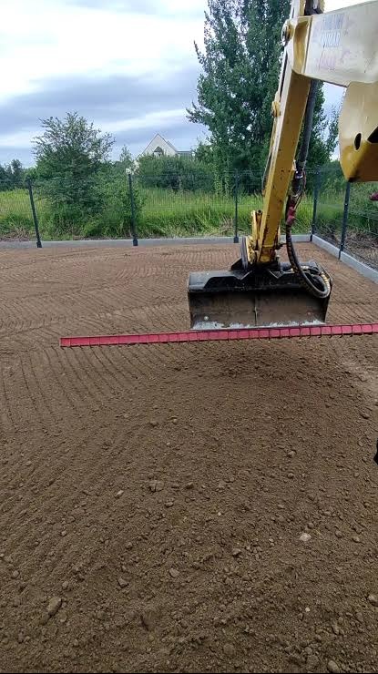 Żółta koparka wyrównuje teren pod budowę, widoczna czerwona listwa poziomująca. W tle ogrodzenie z siatki i dom w budowie. Prace ziemne w toku.