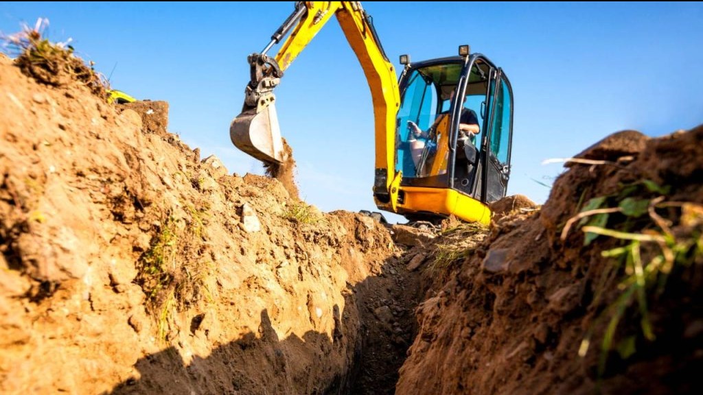 Żółta minikoparka w trakcie pracy, z bliska. Widoczny wykop w ziemi i operator w kabinie. Błękitne niebo w tle. Ujęcie z dołu, podkreślające głębokość wykopu.