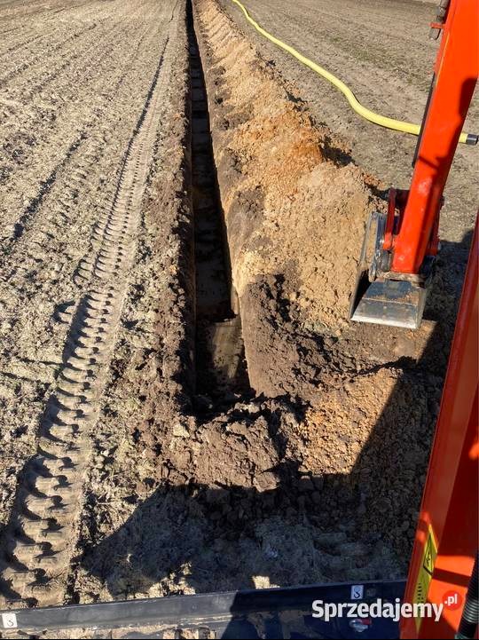 Wąski wykop w ziemi, wykonany równo, z widocznymi śladami opon i fragmentem pomarańczowej maszyny budowlanej. Żółty wąż leży na powierzchni ziemi.