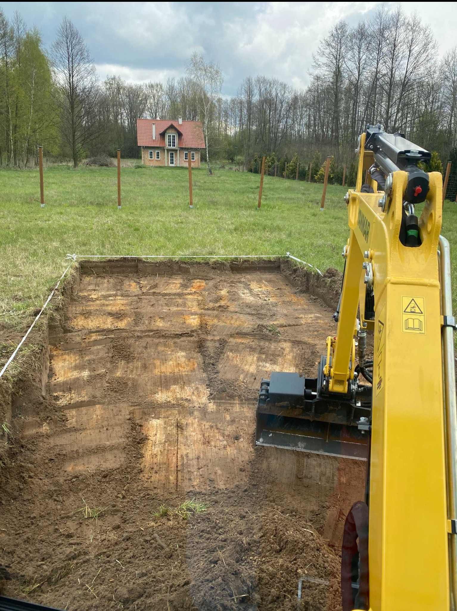Żółta mini koparka Yanmar w trakcie prac ziemnych, widoczny wykop oznaczony sznurkiem, w tle dom jednorodzinny i słupki ogrodzeniowe.