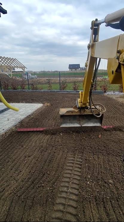 Żółta koparka wyrównuje teren podłoża, widoczne ślady opon i częściowo ułożona kostka brukowa. W tle dom w budowie i ogrodzenie.