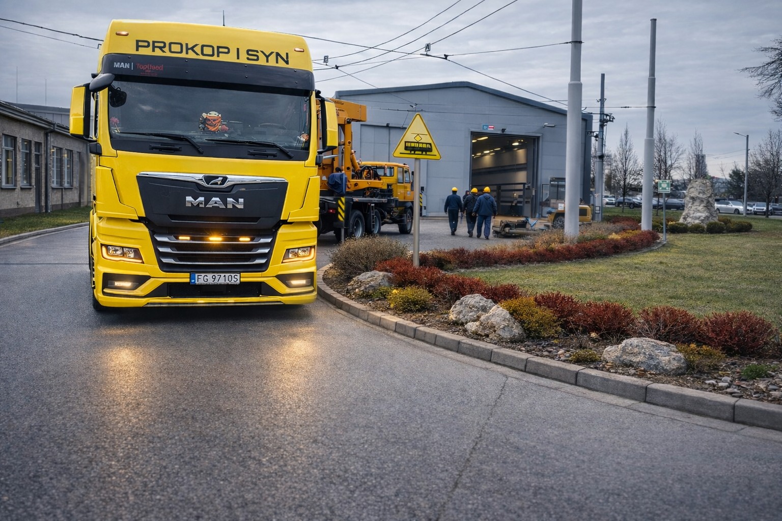Żółta ciężarówka MAN z napisem 'PROKOP I SYN' na tle szarego budynku i grupy robotników w kaskach. Widoczny znak ostrzegawczy i elementy krajobrazu.