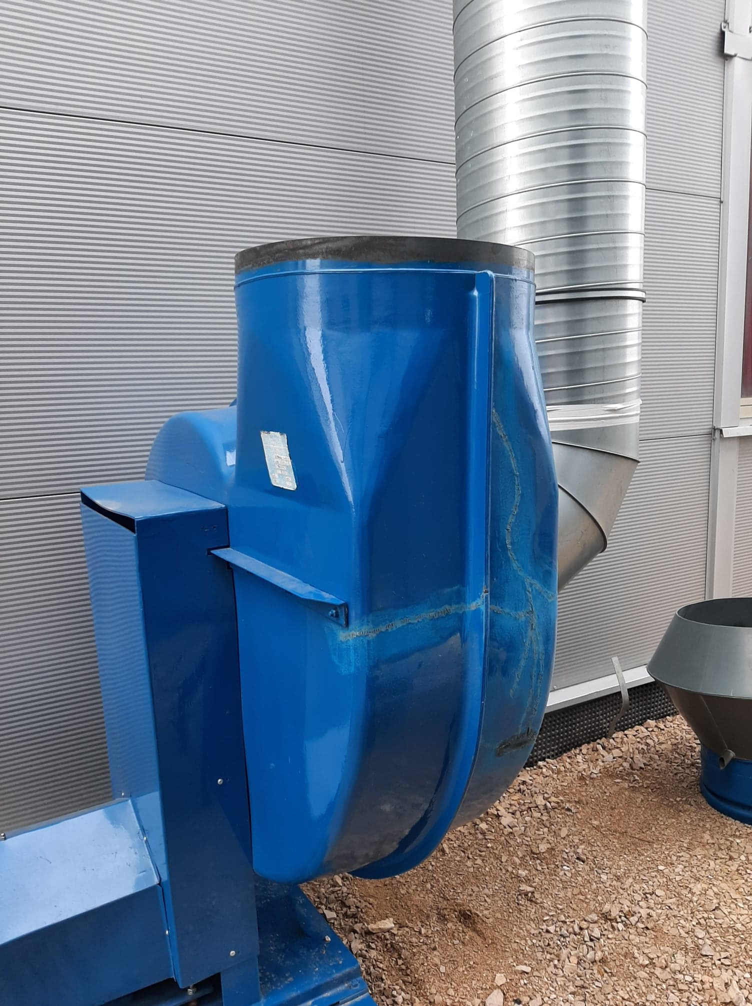 Niebieski wentylator przemysłowy z widocznymi spawami i rurą wentylacyjną na zewnątrz budynku o szarej, blaszanej elewacji.
