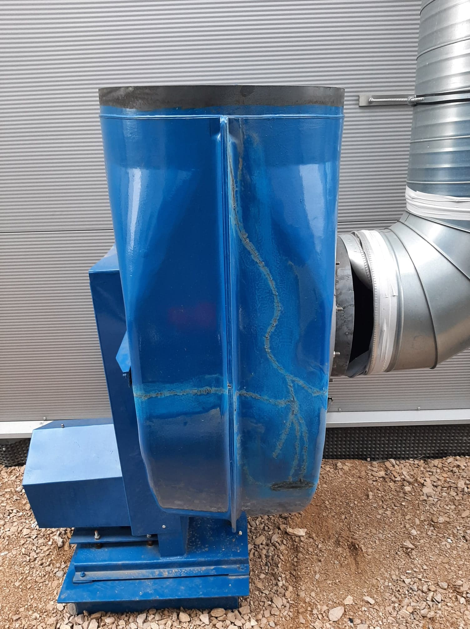 Niebieska maszyna przemysłowa z metalową rurą wentylacyjną, widoczne ślady napraw i spawania, na tle szarej ściany i żwirowego podłoża. Detal złącza.