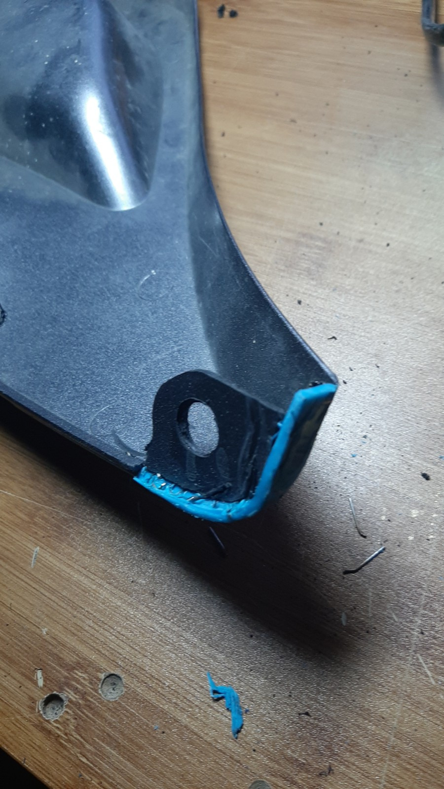 Naprawiony element z tworzywa sztucznego z widocznym spawem, wzmocniony niebieskim tworzywem. Detal z otworem montażowym na drewnianym blacie.