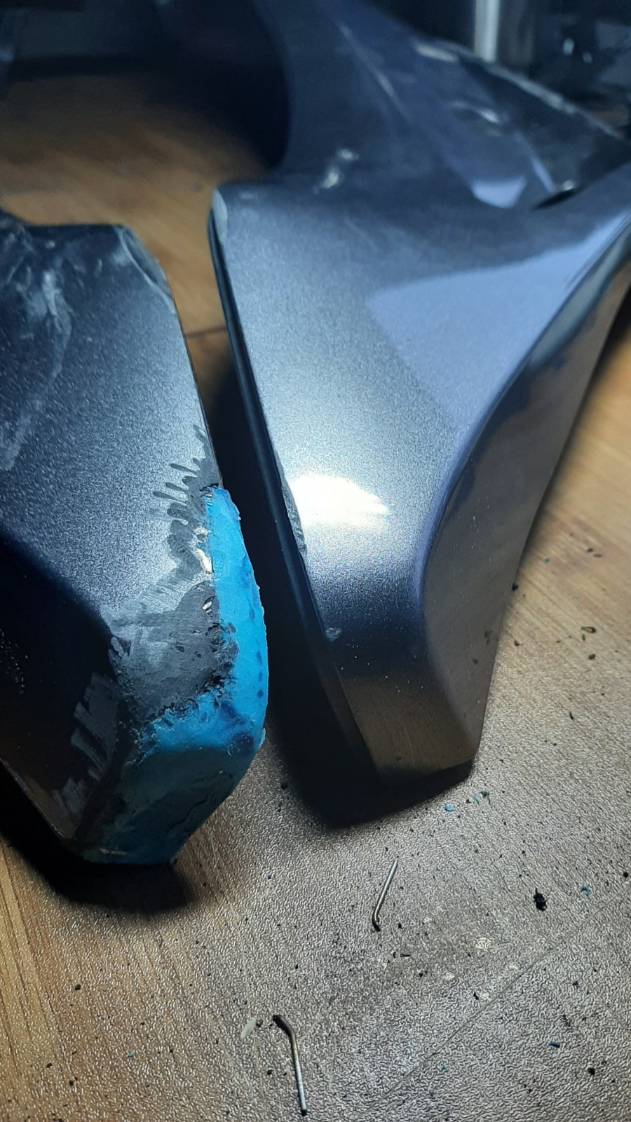 Szary element z tworzywa sztucznego po naprawie, z widocznym niebieskim wypełnieniem ubytku. Dwa metalowe zszywki leżą obok na drewnianej powierzchni.