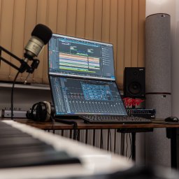 Miłosz Tuszyński - Baga Sound - Profesjonalne stanowisko do miksowania dźwięku z dwoma monitorami, mikrofonem pojemnościowym i klawiaturą MIDI. Widoczne głośniki studyjne i panele akustyczne.
